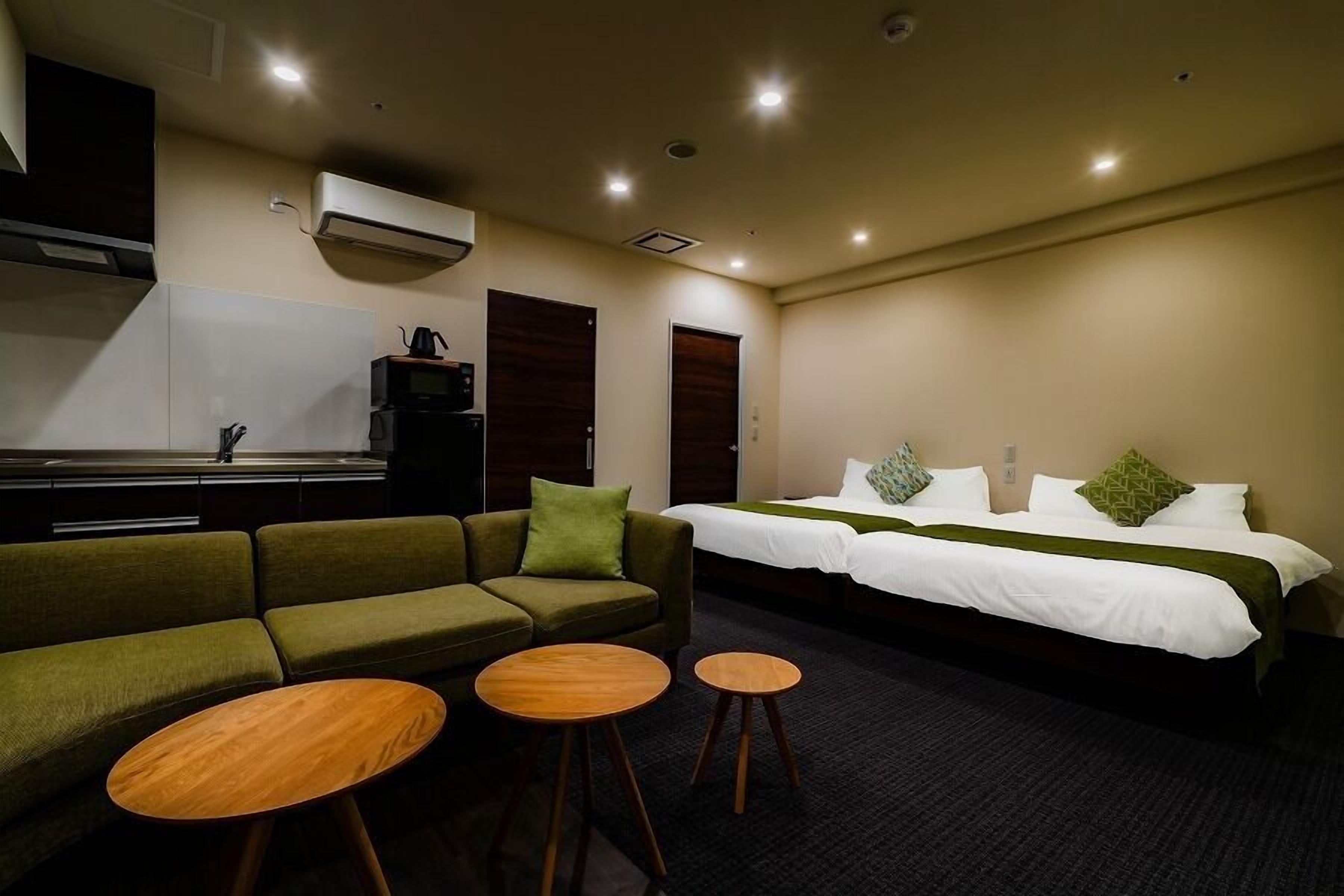 Una habitación de hotel con sofá, dos mesitas, cama con ropa blanca y kitchenette.