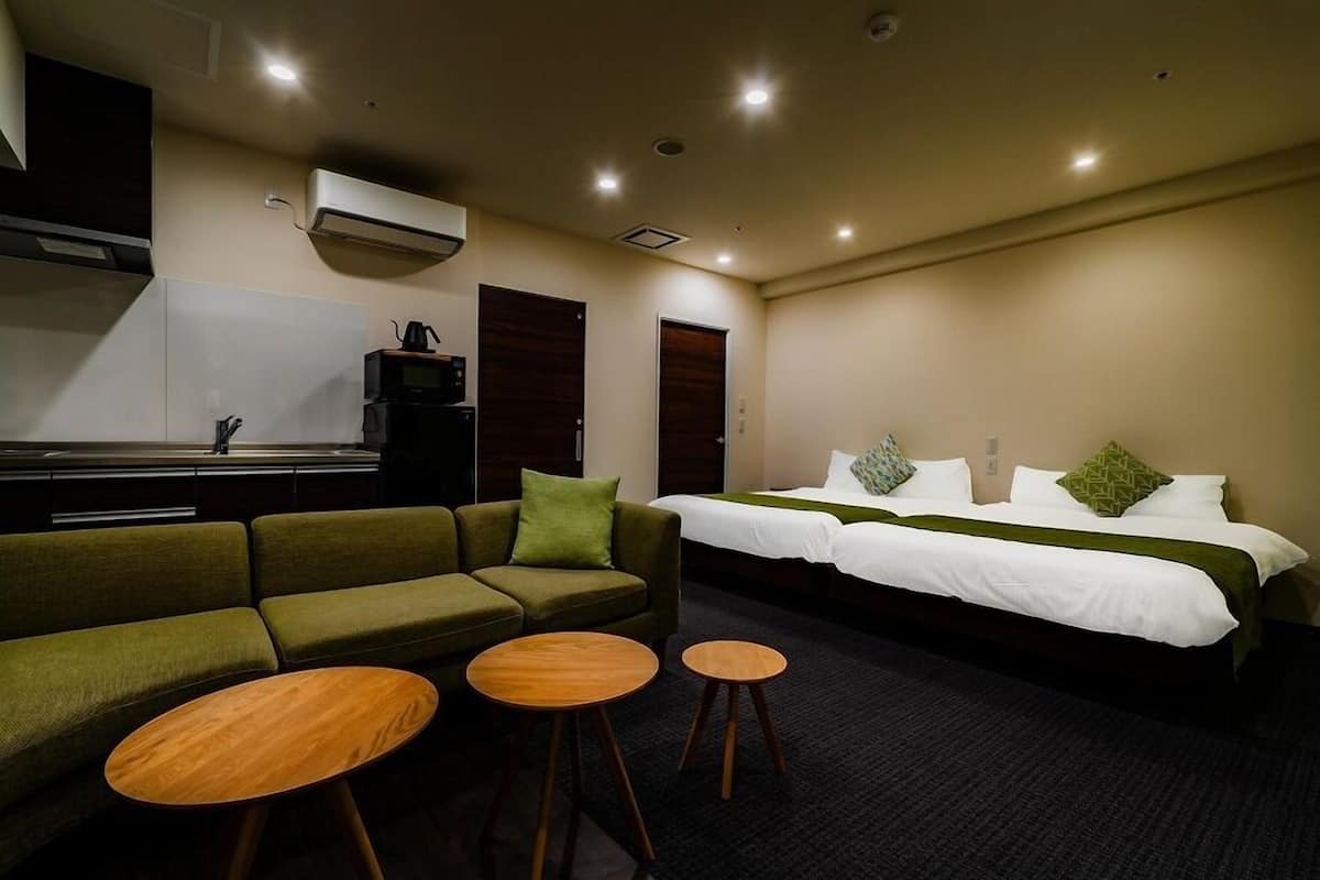 A hotel room with a sofa, two small tables, a bed with white bedding, and a kitchenette.