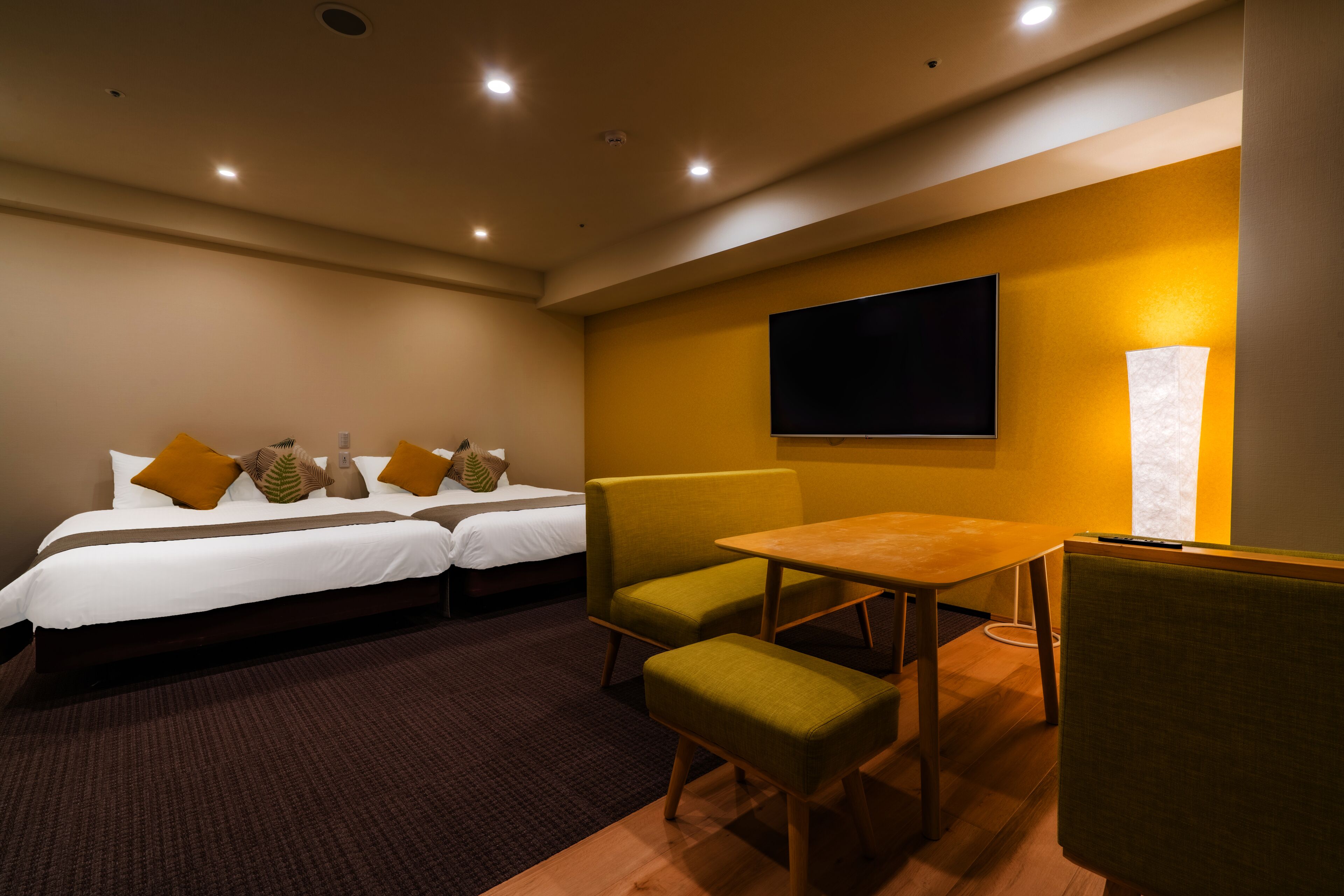 A hotel room with two beds, a dining table, and a TV mounted on the wall.