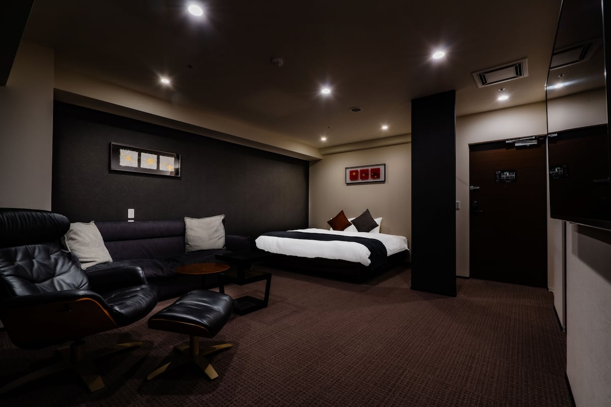 A modern hotel room with a dark-colored sofa, a bed with white bedding, a small table, and a TV mounted on the wall.
