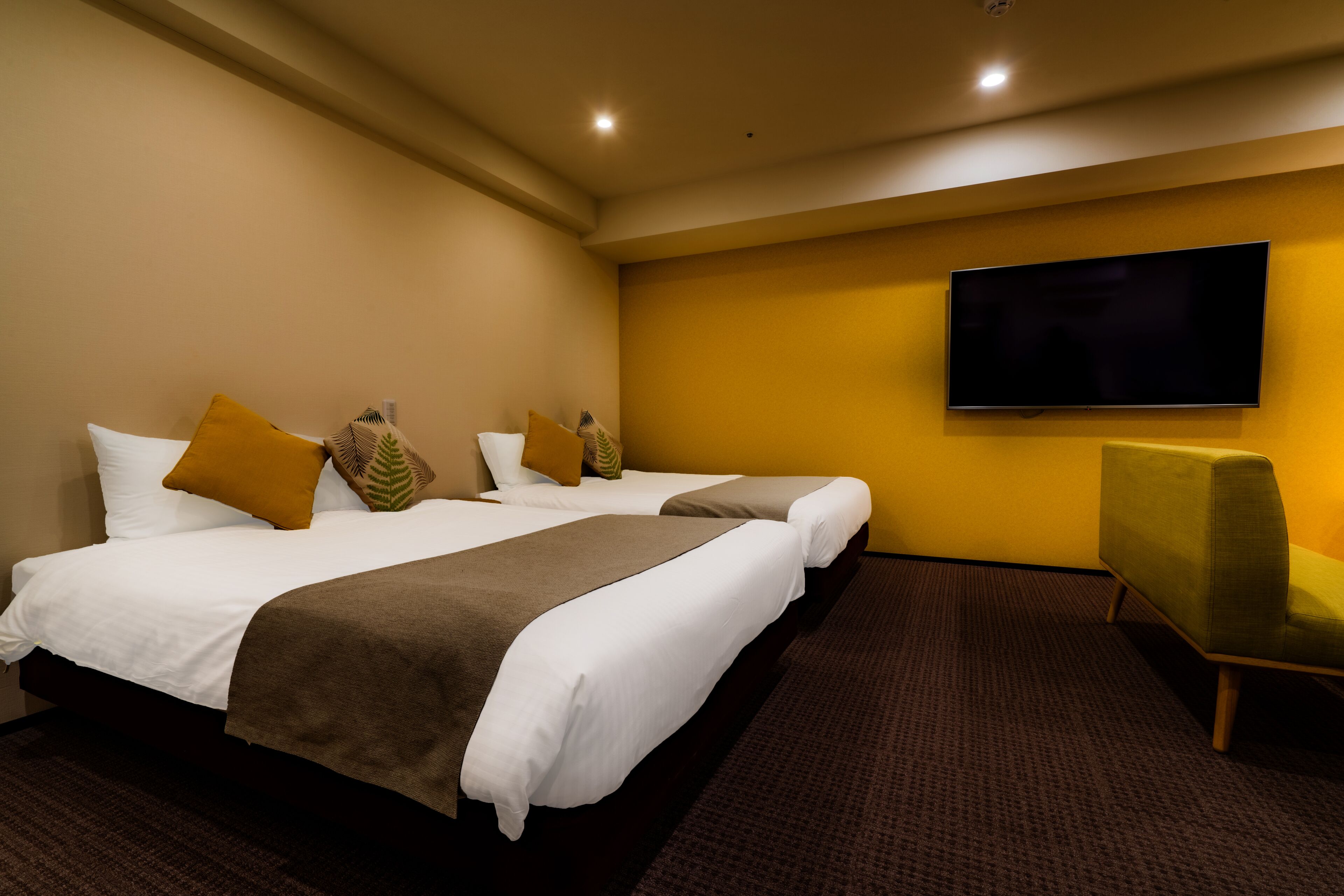 A hotel room with two beds, a flat-screen TV, and a green armchair.