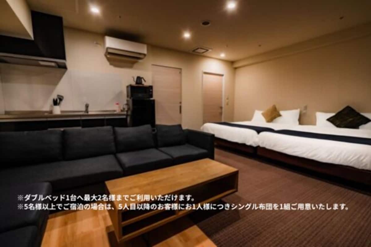 A hotel room with a sofa, two single beds, a small table, and a kitchenette.