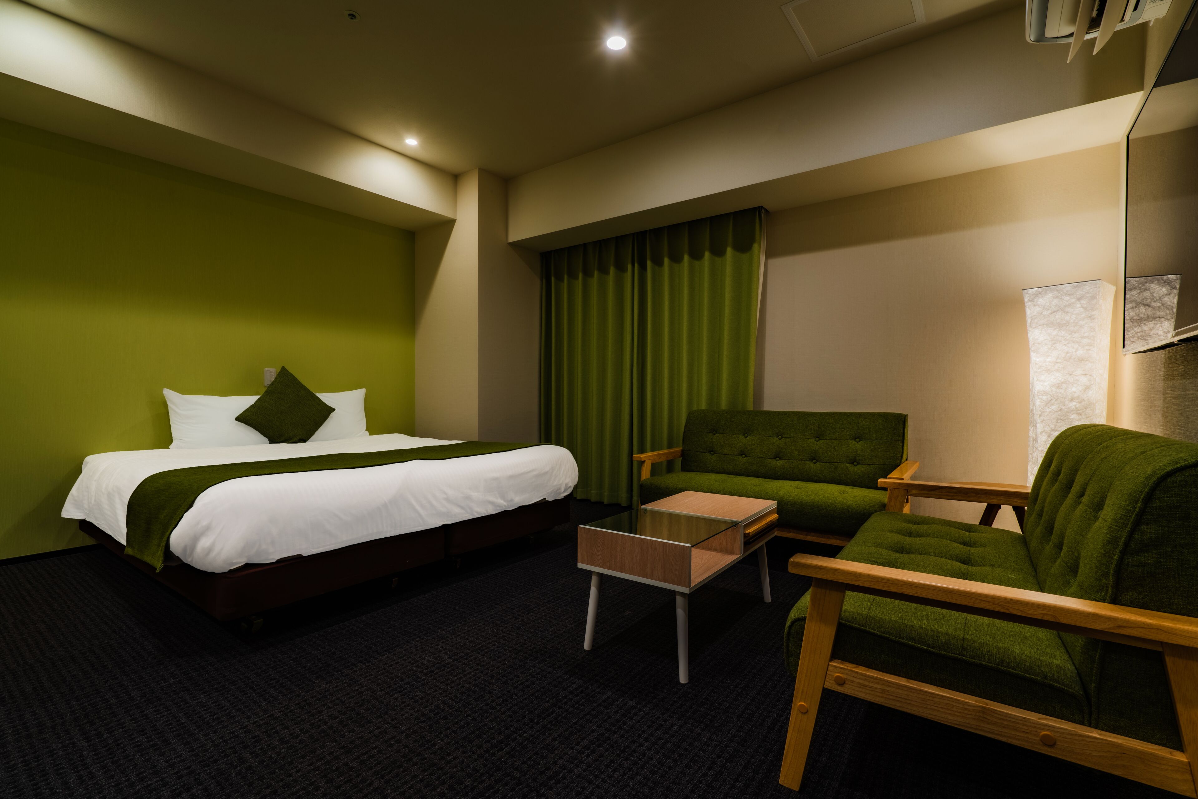 A hotel room with a bed, a green armchair, a wooden side table, and a green curtain.
