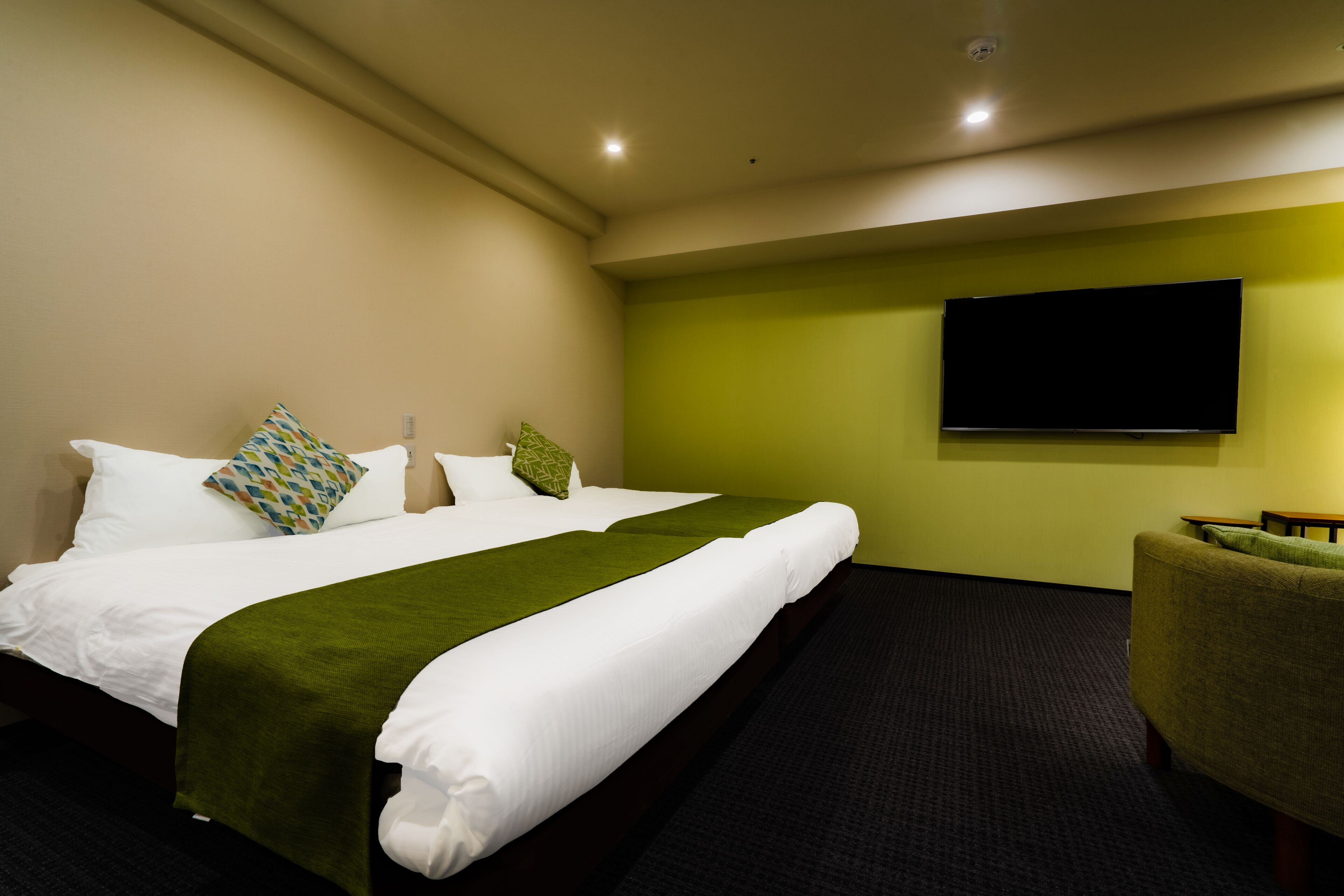 A hotel room with a bed, green wall, and a flat-screen TV mounted above it.