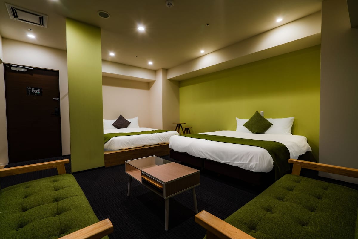 A hotel room with two beds, a small table, and green upholstered chairs.