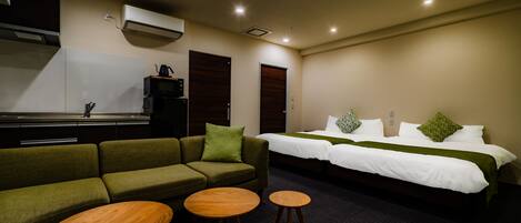 Een hotelkamer met een groene bank, twee houten tafels, een bed met wit beddengoed en een kitchenette op de achtergrond.