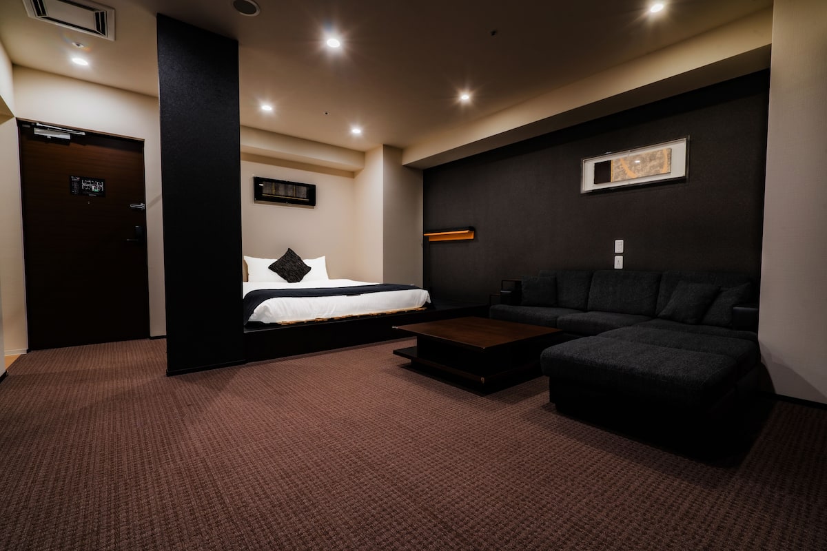A modern hotel room with a bed, a sofa, a coffee table, and a dark wall with a framed picture.