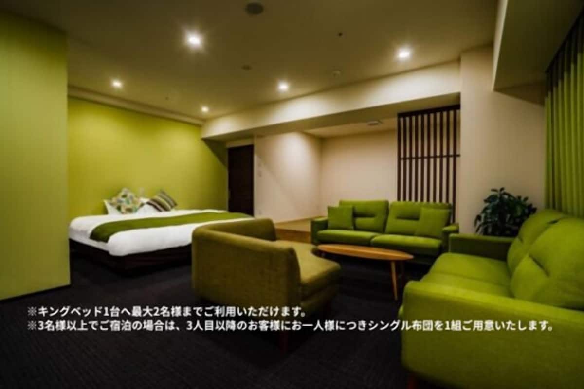 A hotel room with a bed, two green armchairs, a small table, and a green sofa.