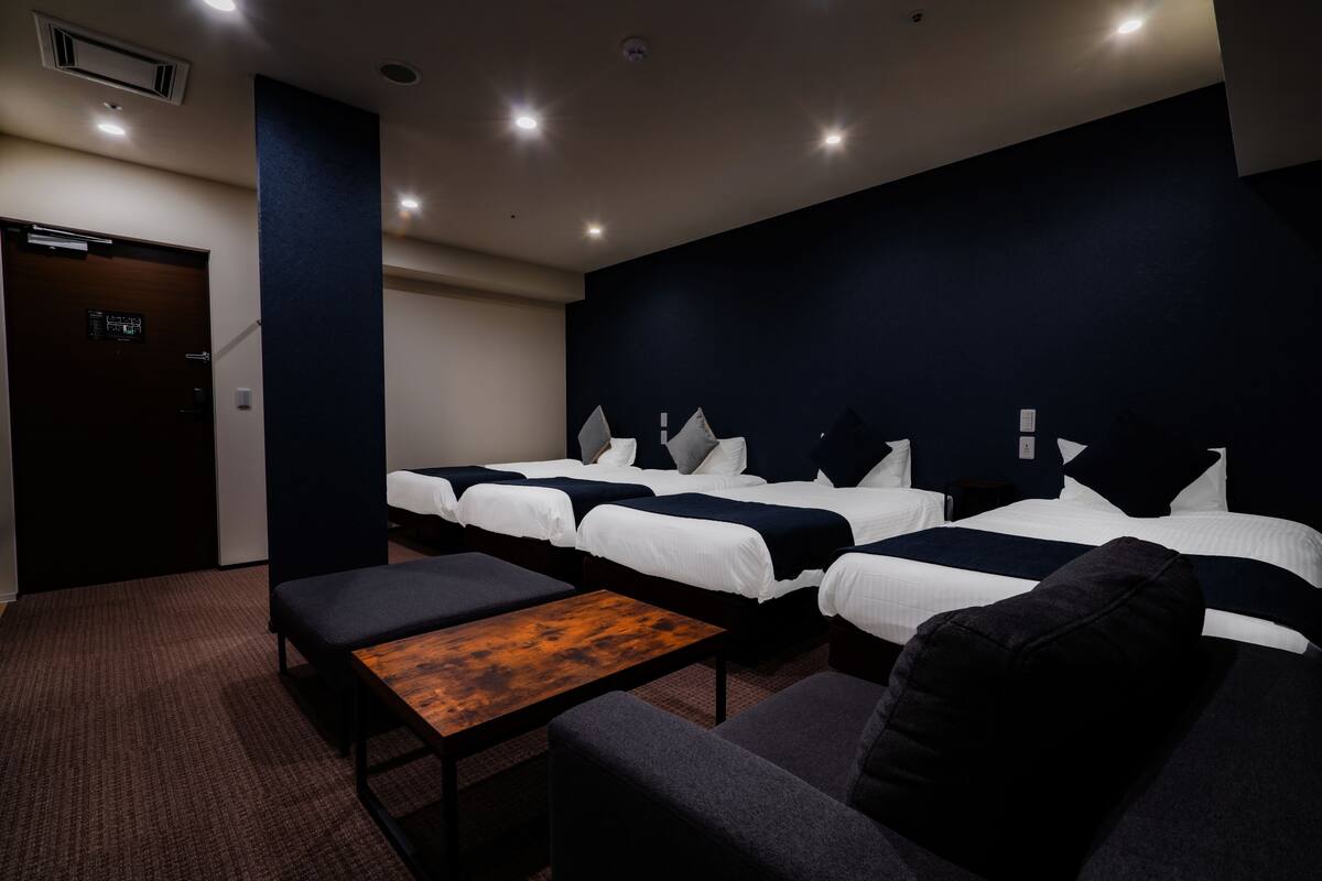 A hotel room with three beds, a small table, and a bench.