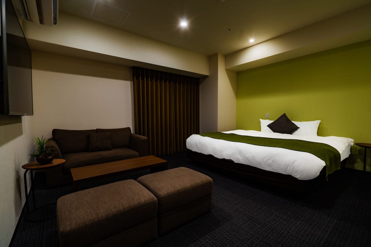 A hotel room with a bed, a sofa, an ottoman, a small table with a plant, and a wall-mounted TV.