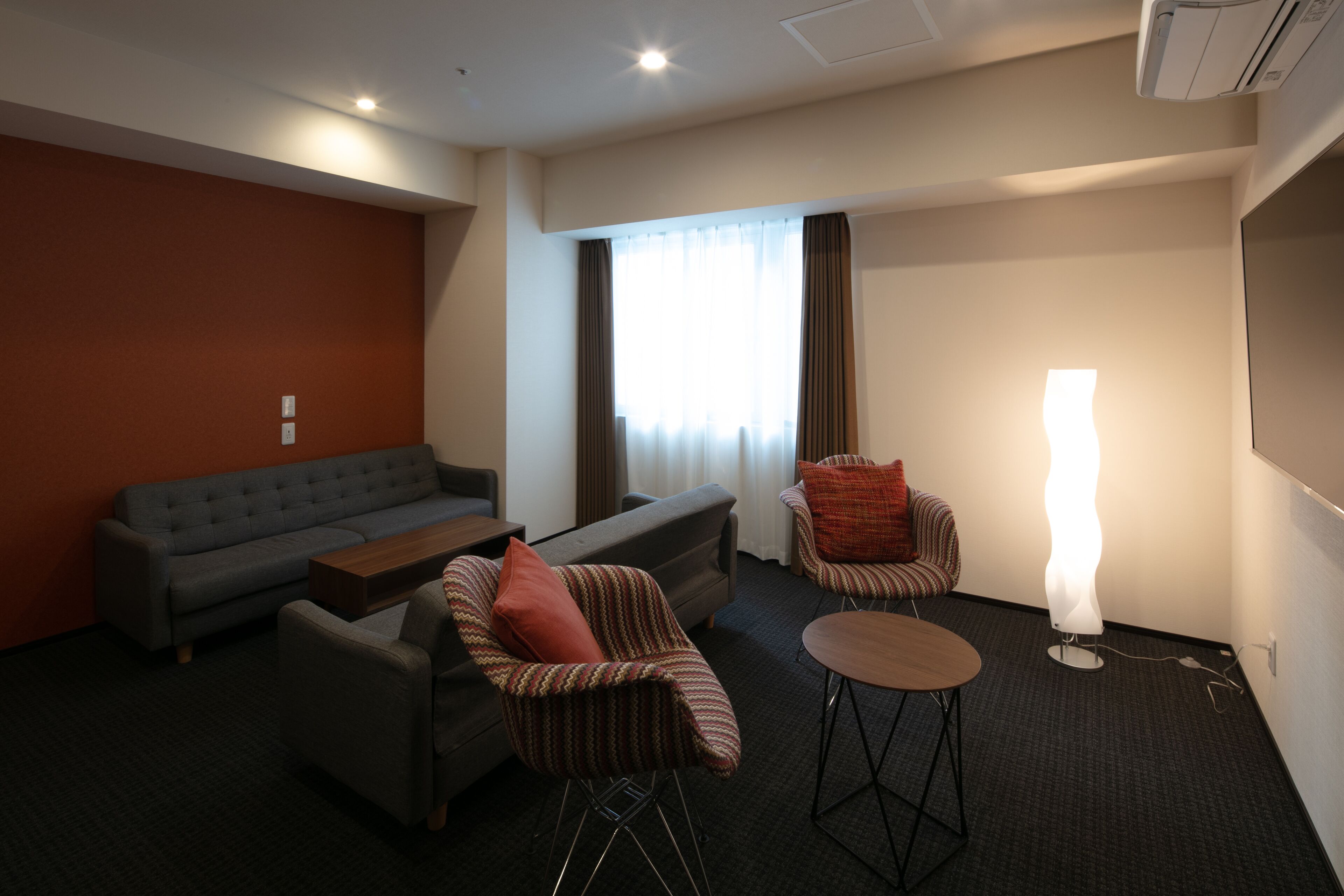 A modern hotel room with a grey sofa, striped armchairs, a small round table, and a standing lamp.