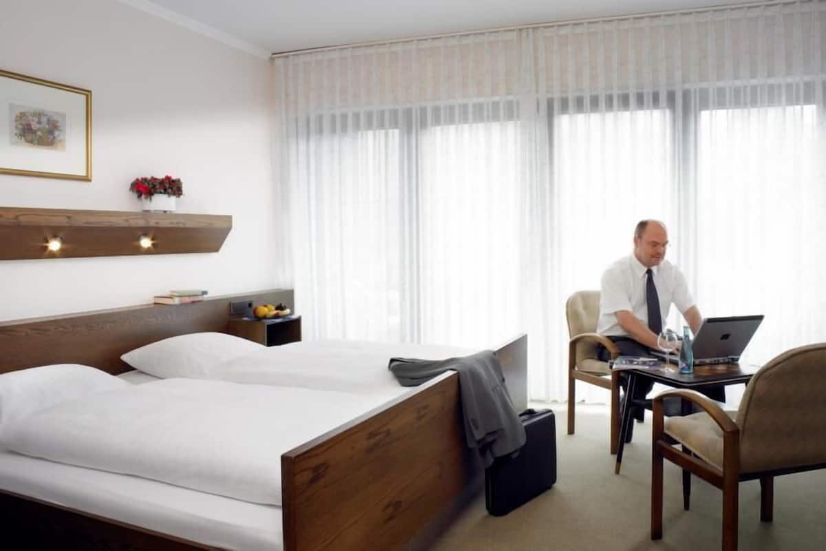 Quarto de hotel com uma cama, uma escrivaninha e uma cadeira. Há uma janela com cortinas leves, uma estante com livros e um vaso de flores, e um quadro na parede.