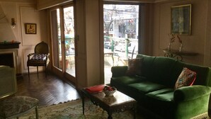 A living room with a green sofa, a wooden coffee table, a patterned rug, and a chair. There is a fireplace, a painting on the wall, and a view of the street outside. - My home in Buenos Aires | 1937 sqft | 2 bdr | n/STelmo (Barracas)