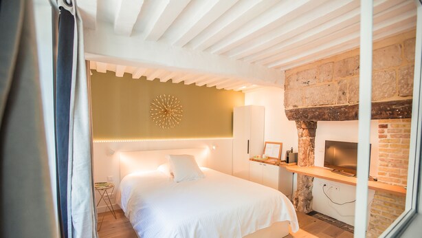 A bedroom with a bed, a wall-mounted light fixture, a brick fireplace, and a small table with a picture frame. - La Maison des Arts (Honfleur)