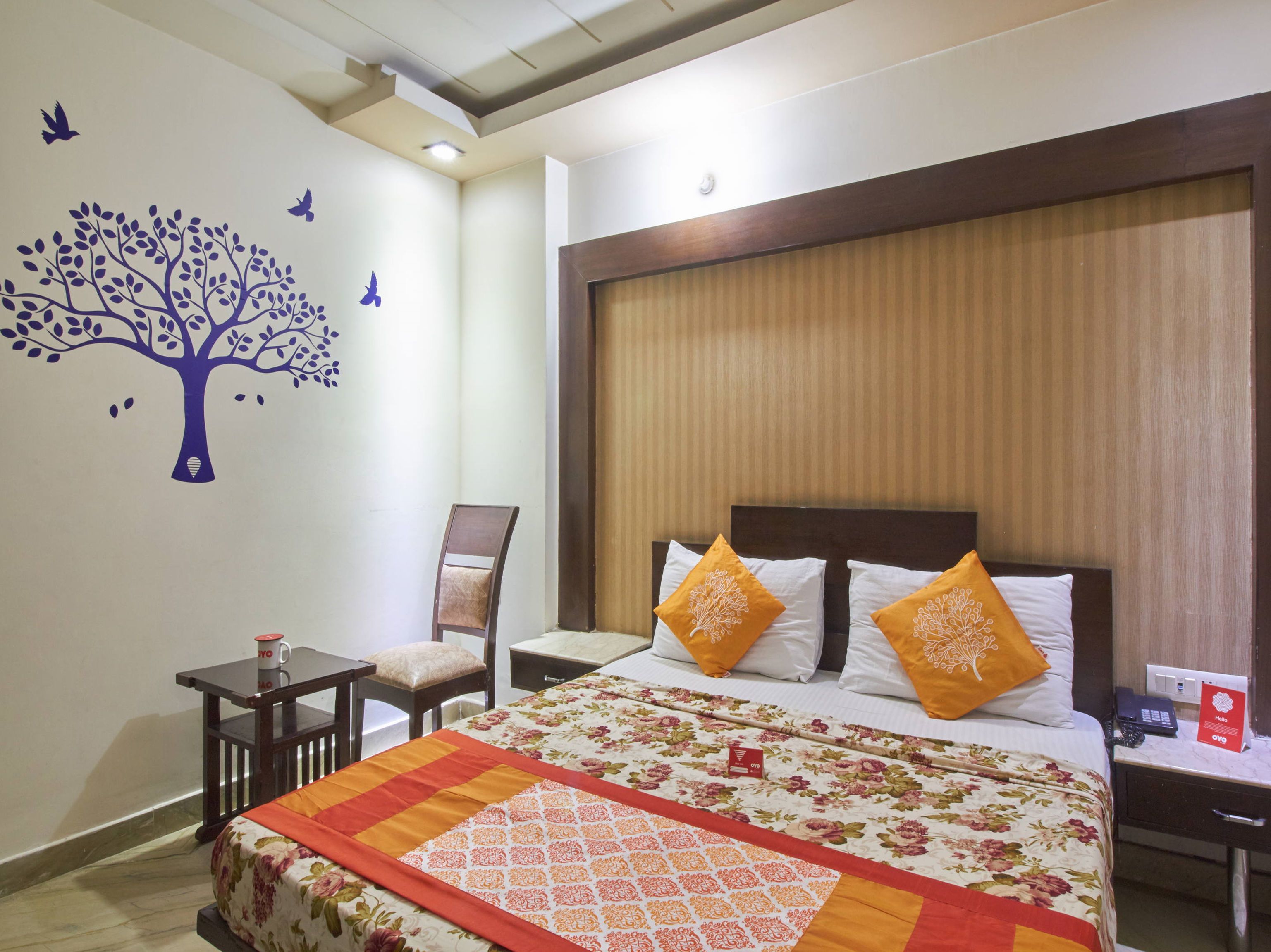 A hotel room with a large bed, two pillows, a nightstand, a chair, a small table, and a wall decoration of a tree with birds.