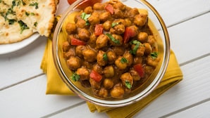 Een kom kikkererwten curry met groenten, vergezeld van naanbrood en een tomaat op een witte houten tafel.