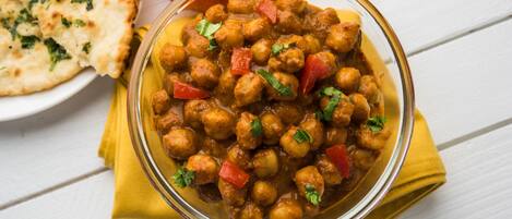 A bowl of chickpea curry with vegetables, accompanied by naan bread and a tomato on a white wooden table.