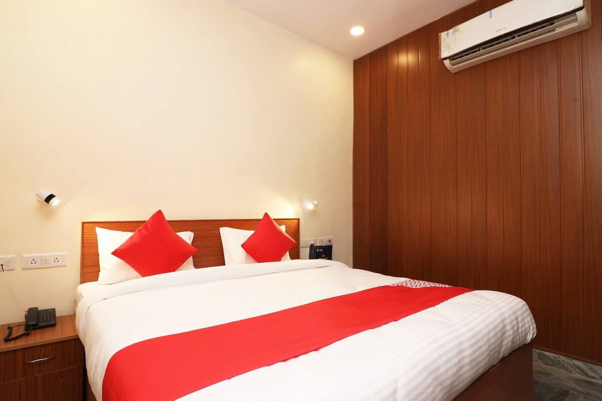 Un lit bien fait, avec une literie blanche rehaussée de touches de rouge, une tête de lit en bois et un appareil de climatisation fixé au mur.