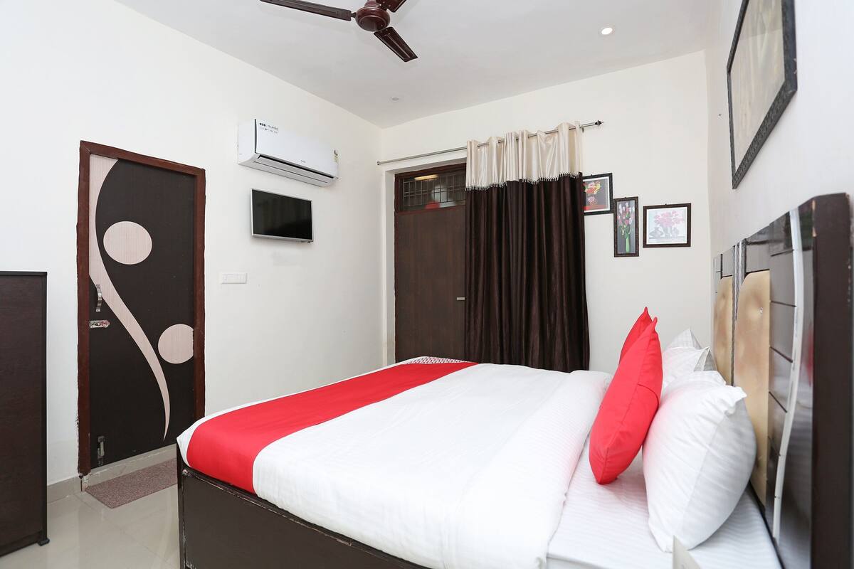 A hotel room with a bed, a ceiling fan, an air conditioning unit, a door, and a window with curtains.