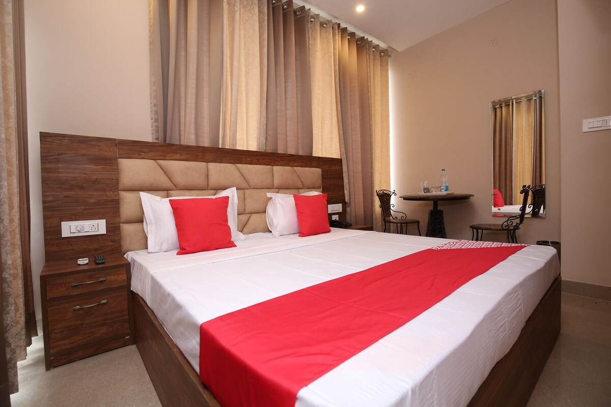 A hotel room with a large bed, a wooden nightstand, a small table with a chair, and a mirror on the wall.