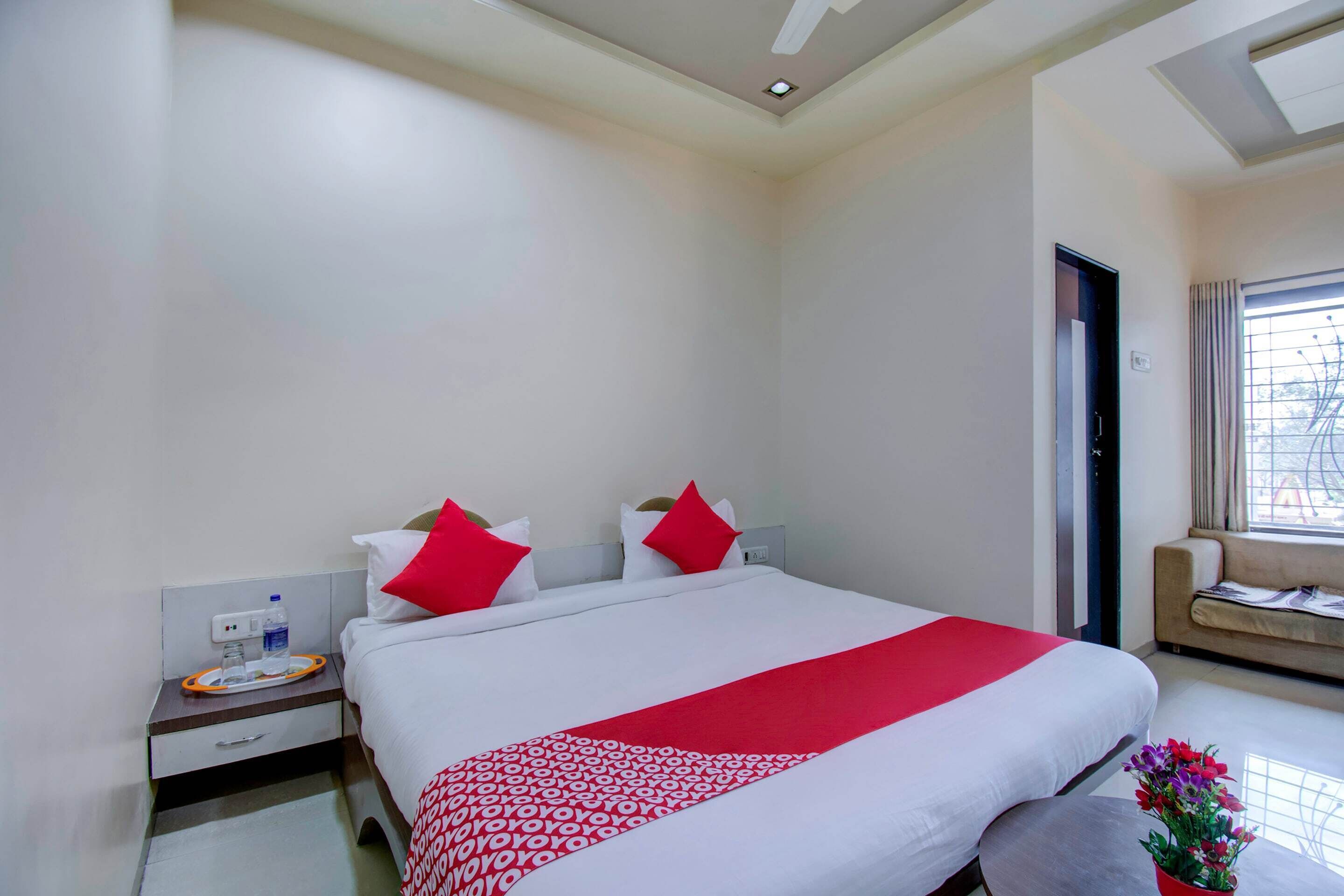 A hotel room with a large bed, a bedside table with a water bottle, a red throw, and a window with a view.