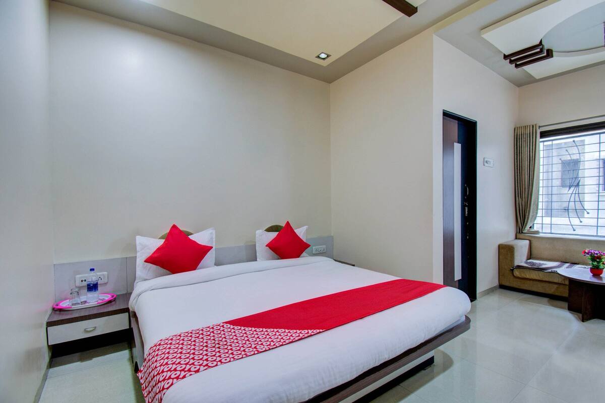 Habitación de hotel con una cama grande, dos almohadas rojas, una mesita de noche con una botella de agua y una ventana con cortinas.