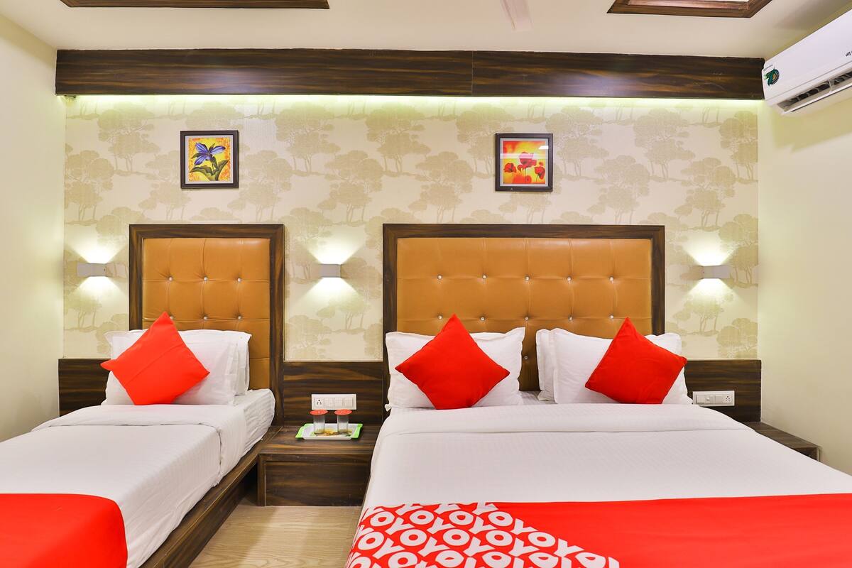 A hotel room with two beds, each with a wooden headboard and red pillows. There are wall-mounted lights and two framed pictures on the wall.