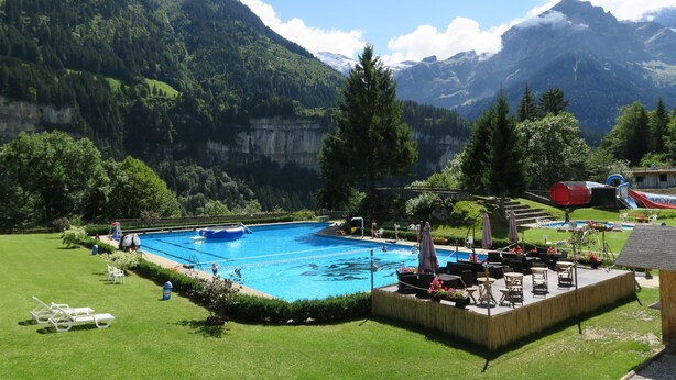 A swimming pool with a deck area, surrounded by a lush green landscape and mountains in the background. - Hotel le Palladium (Champery)