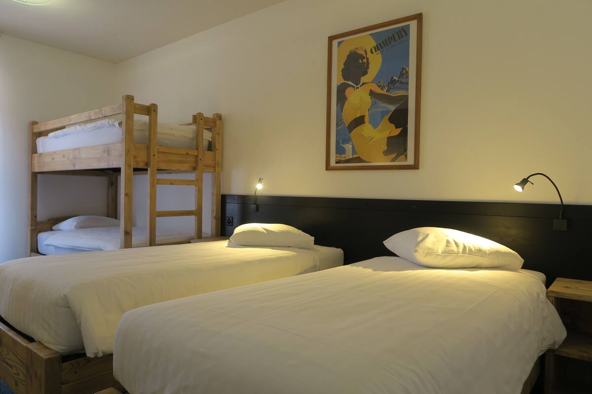 Una habitación con dos camas individuales, una litera, un cuadro enmarcado en la pared y una mesita de noche con lámpara.