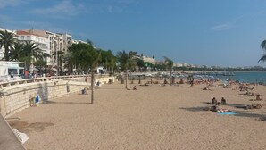 Een strand met mensen die zonnebaden en wandelen. Er staan palmbomen en een gebouw op de achtergrond.