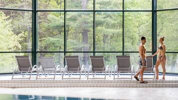 Una piscina interior con paredes de vidrio transparente, rodeada de sillas de descanso y vistas a una zona boscosa.