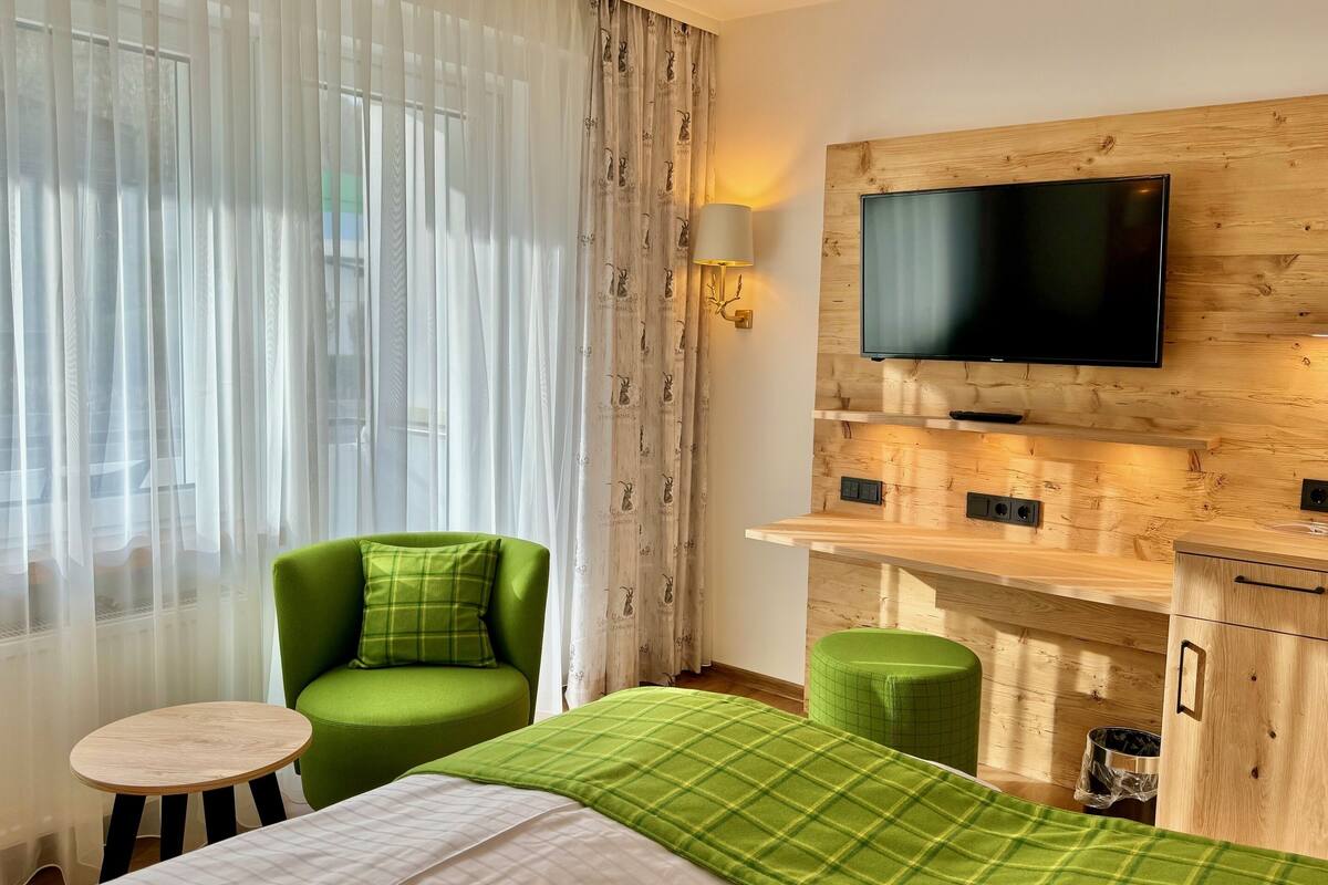 Habitación de hotel con una cama, un sillón verde, una mesita redonda pequeña, un reposapiés verde, un televisor montado en una pared de madera y una ventana con cortinas traslúcidas.