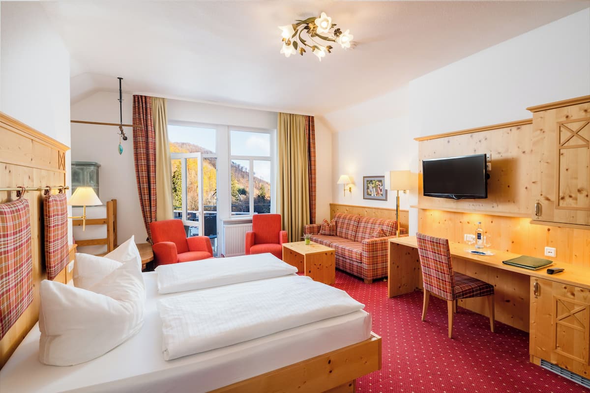 Habitación de hotel con cama, dos sillas rojas, un sofá, un escritorio, televisor y ventana con vista a montañas.