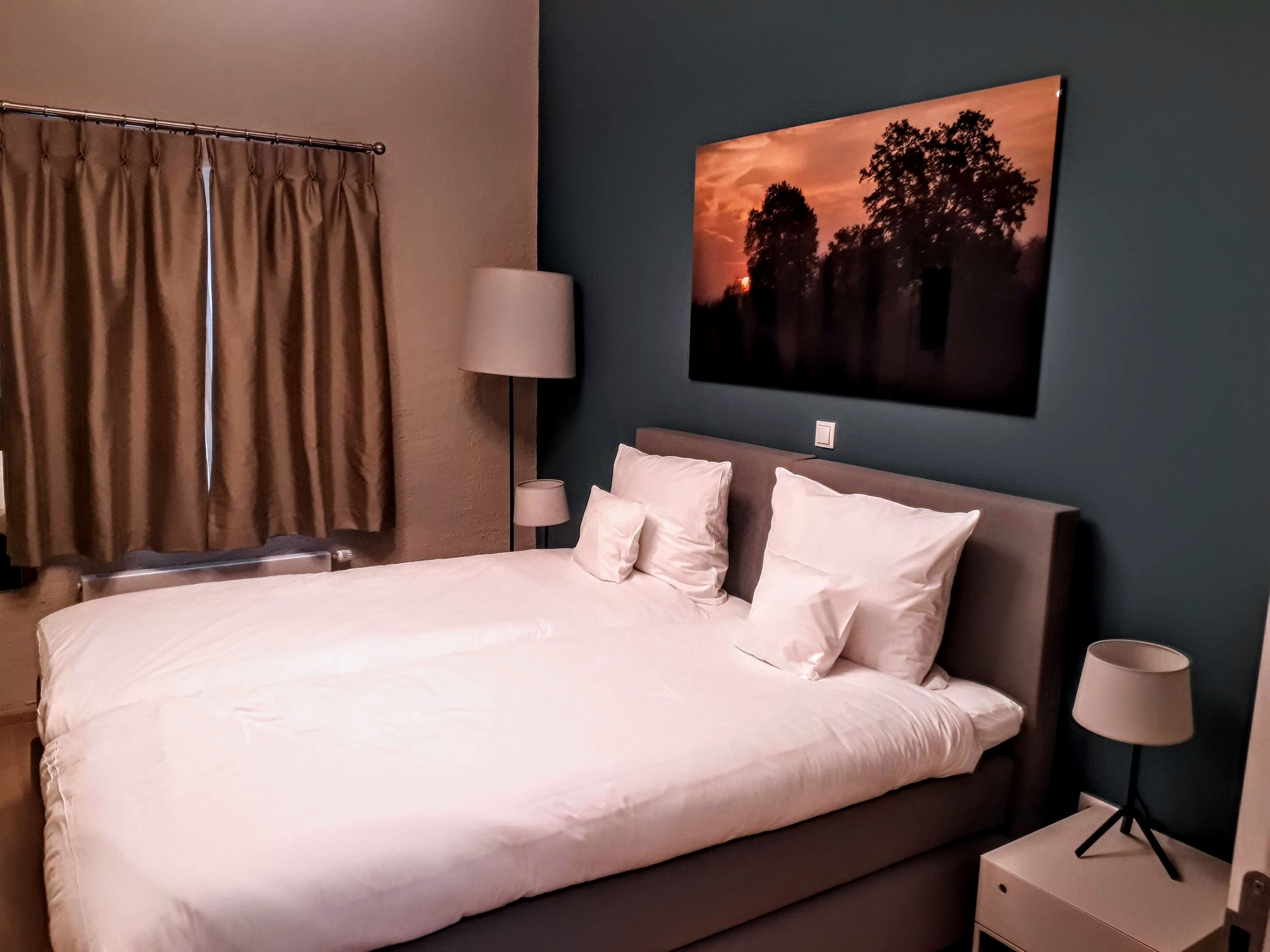 A neatly made bed with white linens and pillows, a bedside lamp, and a wall-mounted picture of a sunset.