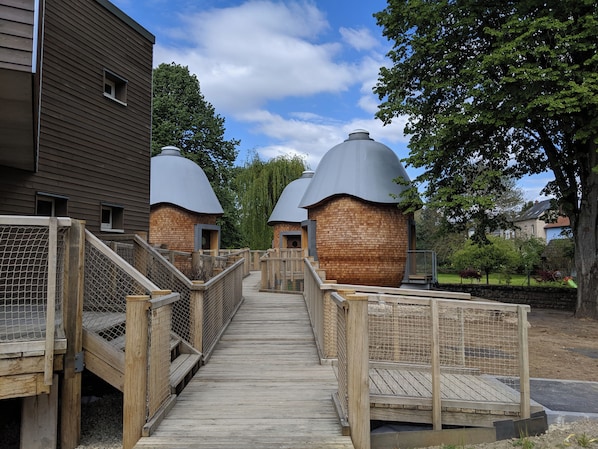 A wooden pathway leading to a row of dome-shaped buildings with a brick base. - HôtelBrasserie & Ecolodges Péitche Lauer (Useldange)