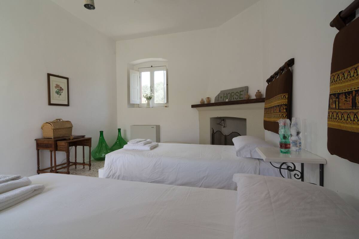 Une chambre d’hôtel avec deux lits, une petite table sur laquelle se trouve un panier, une fenêtre donnant sur un paysage agréable et une cheminée surmontée d’une inscription.
