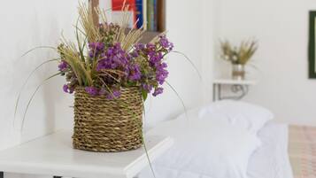 A wicker basket with purple flowers and green foliage on a white table in a room with a bed and a small side table with a plant.