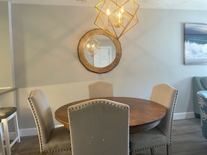 A dining area with a round wooden table, four upholstered chairs, a wooden-framed mirror, and a geometric chandelier.
