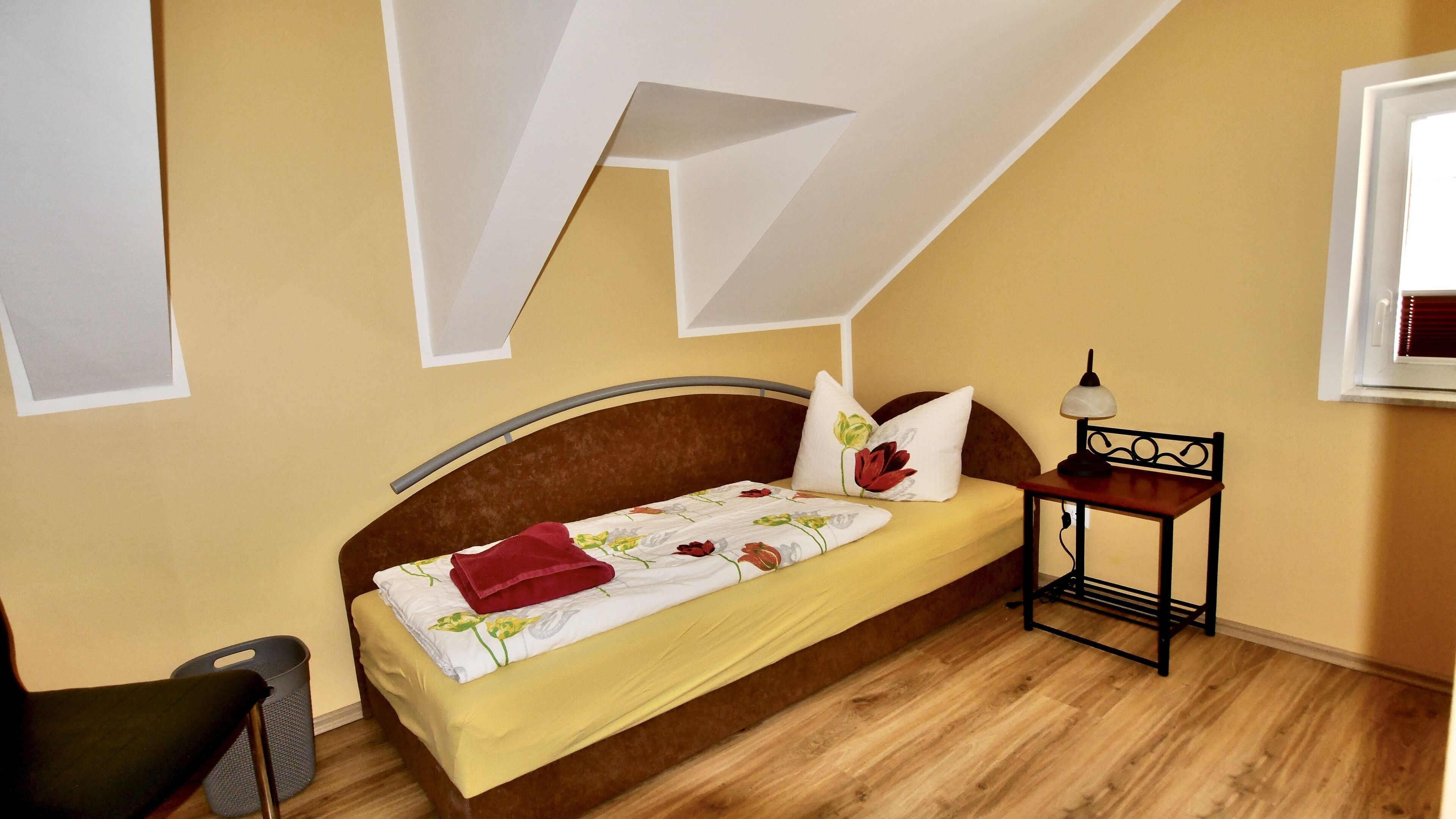 A bedroom with a bed, a bedside table with a lamp, a chair, and a window.