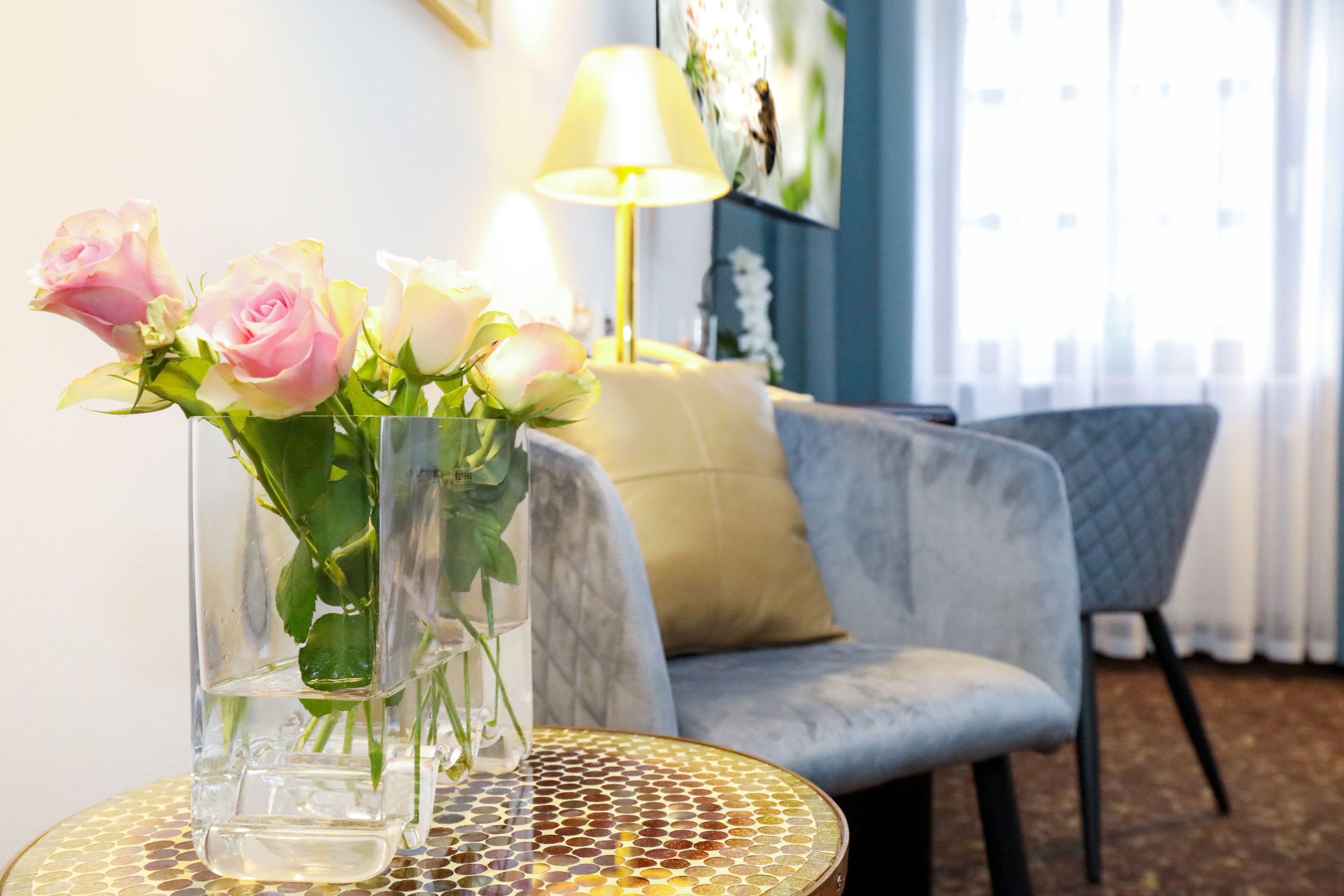Se observa claramente una habitación con una mesa redonda, un florero con rosas rosadas, una lámpara y un sofá gris.