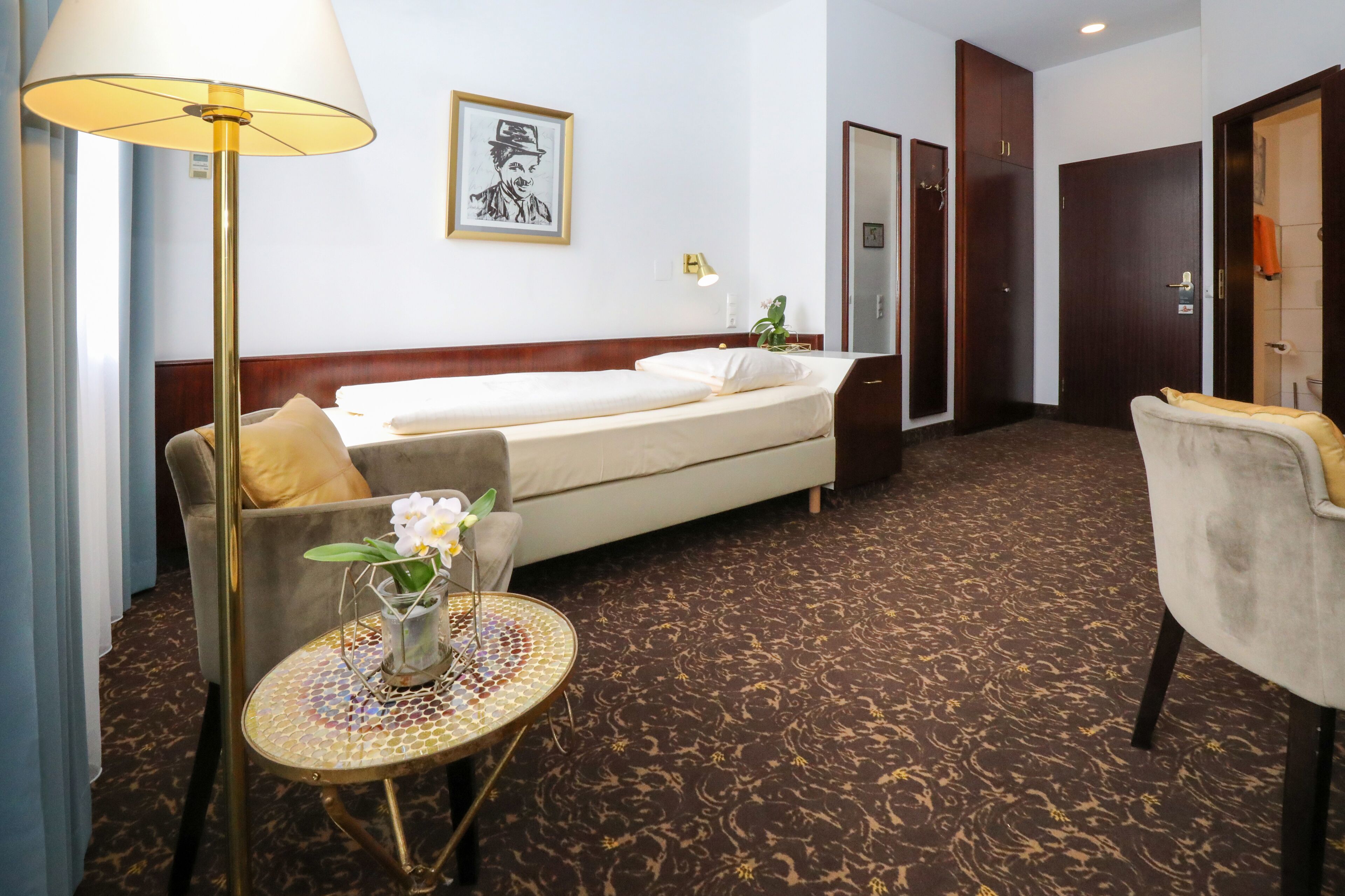 A hotel room with a bed, a sofa, a small table with a vase of flowers, a lamp, and a framed picture on the wall.