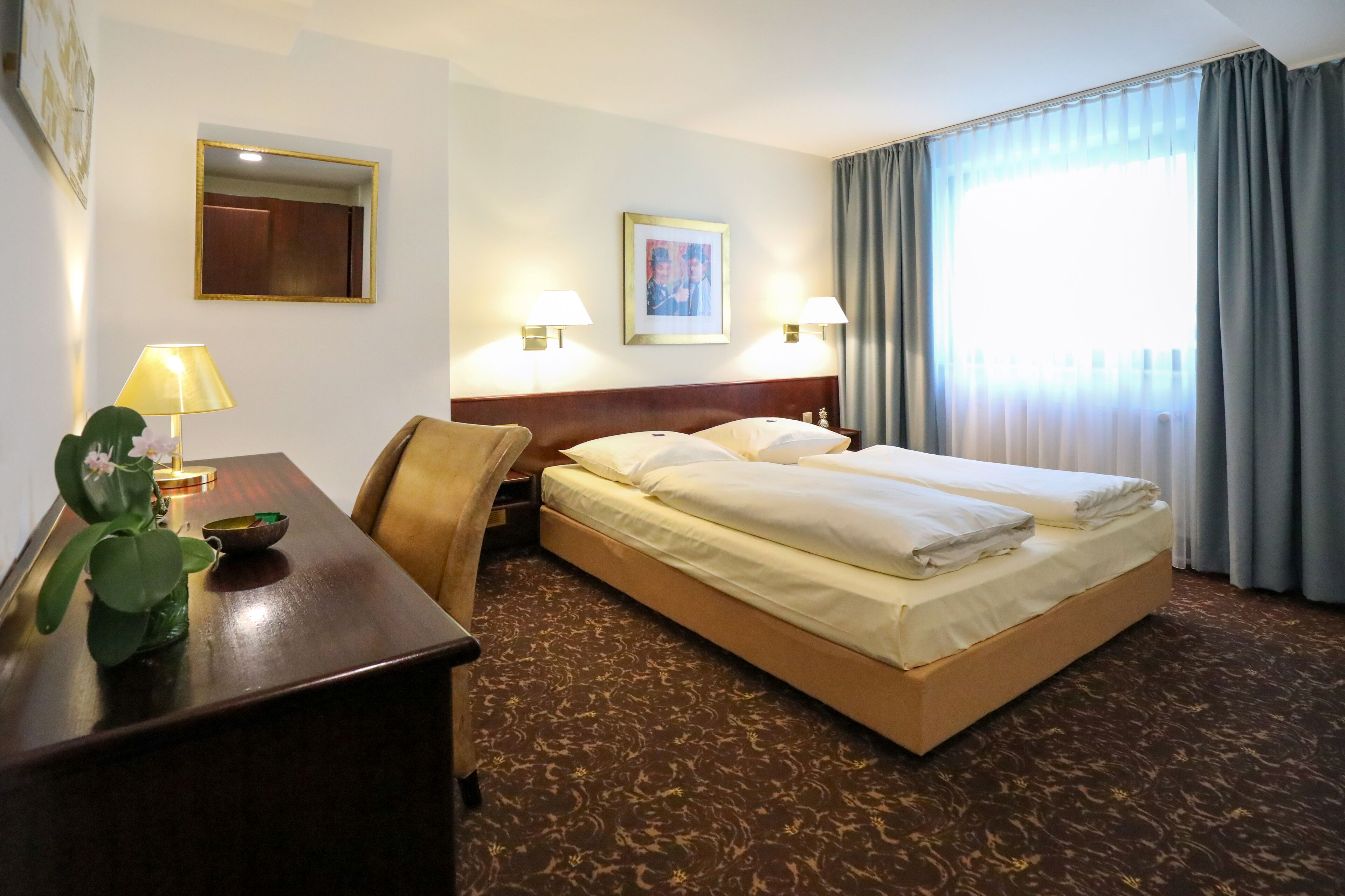 Quarto de hotel com cama, abajures, escrivaninha com luminária, espelho e janela com cortinas.