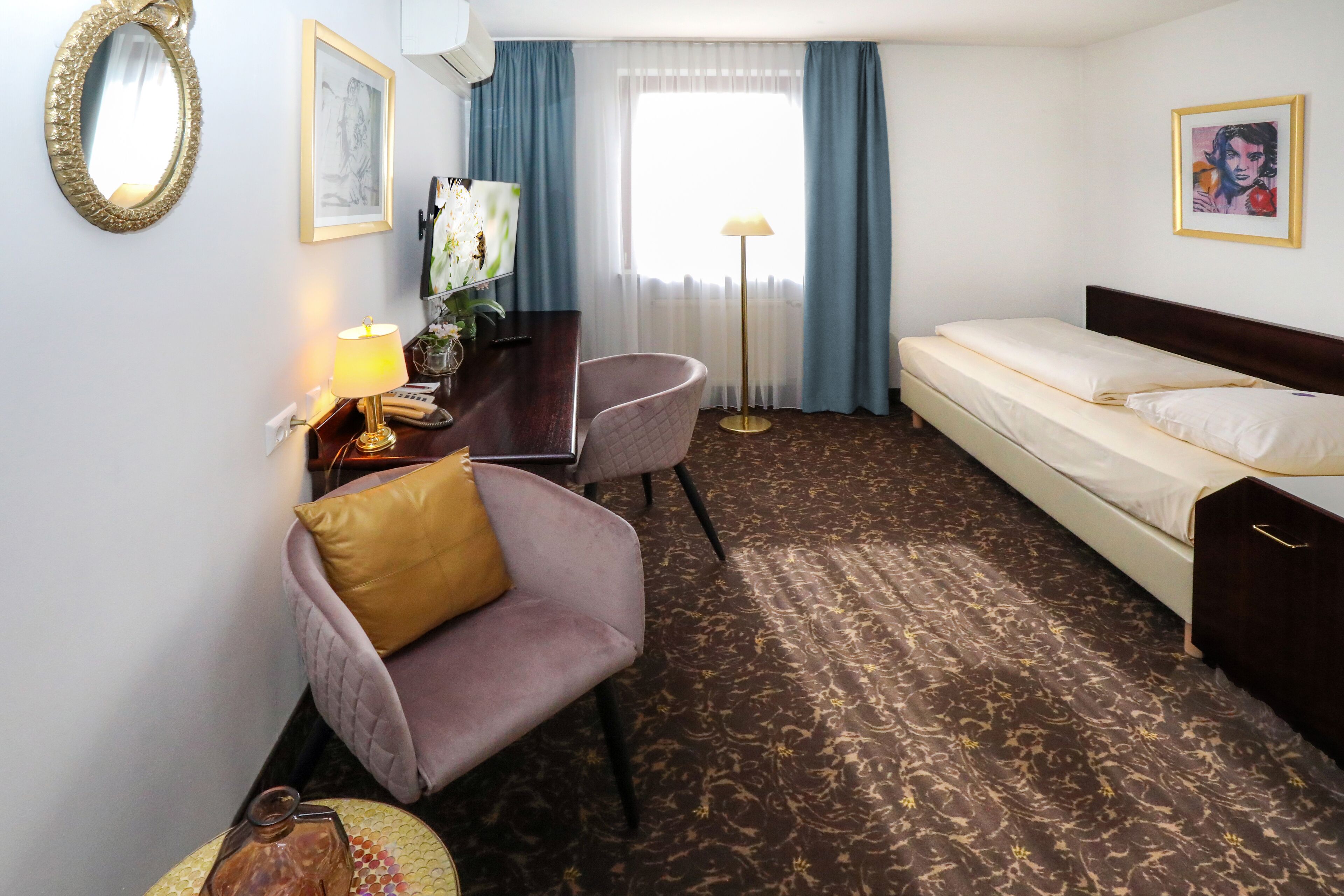 A hotel room with a bed, a desk with a lamp, a chair, a mirror, and a window with curtains.