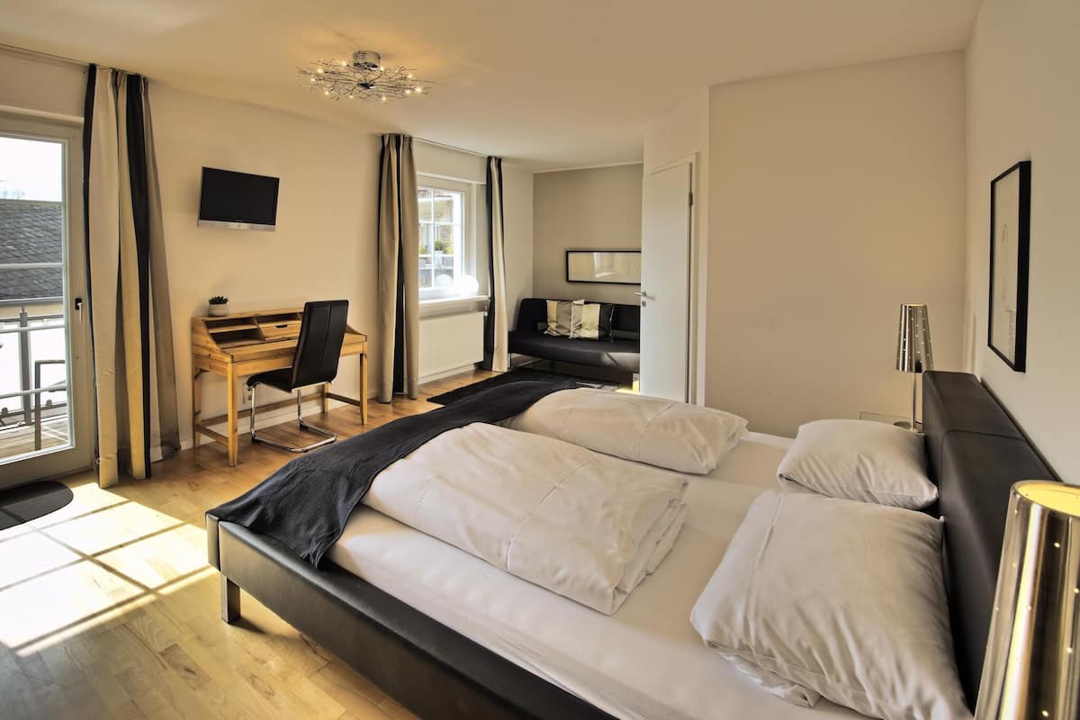 A bedroom with a large bed, a desk, and a chair. There is a TV mounted on the wall, a window with curtains, and a door.