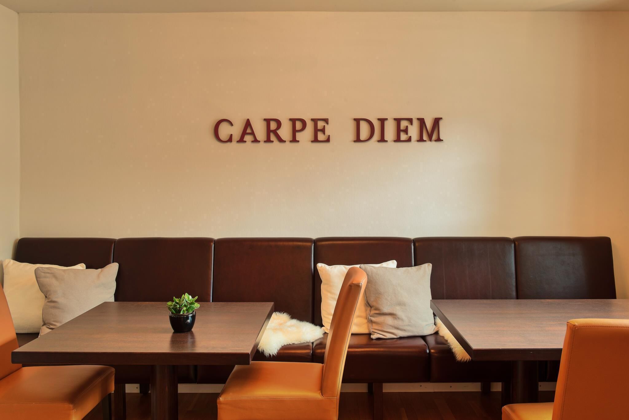 A dining area with a long wooden table, brown leather booth seating, and a wall-mounted sign that reads "CARPE DIEM".