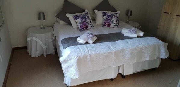 Una cama bien hecha con una colcha blanca y dos almohadas, una mesita de noche con una lámpara y una estantería con otra lámpara.
