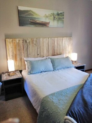 Un dormitorio con cabecera de madera, una cama grande con ropa blanca, dos mesitas de noche con lámparas y un cuadro de un paisaje fluvial sobre la cama.