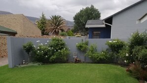 Un césped bien cuidado con un bajo muro de piedra, un pequeño jardín con diversas plantas, y un edificio de fachada gris y tejado azul.