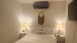 A bedroom with a bed, two bedside tables with lamps, a wall-mounted air conditioner, a television, and a decorative wall hanging.