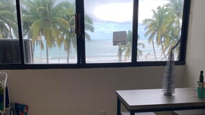 A room with a large window overlooking a beach with palm trees and the ocean. There is a bed with a quilted cover and a yellow pillow, a small table with a decorative item, and a chair.